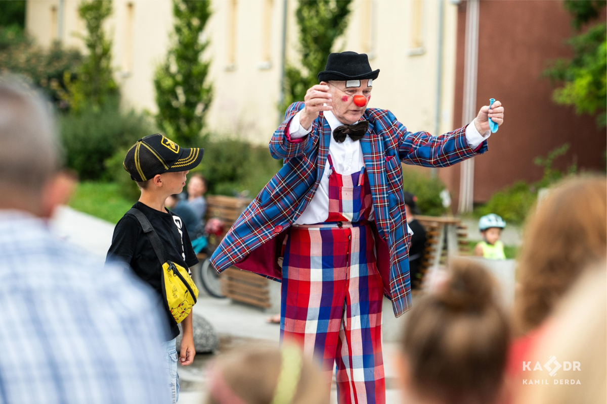 Klown w kraciastej marynarce i meloniku idzie z podniesionymi rękami, obok chłopiec