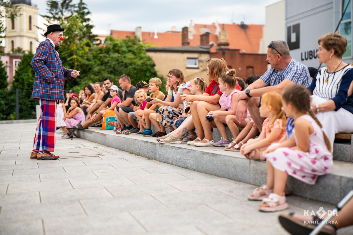 klaun stoi przed dwoma rzędami dzieci na placu teatralnymi i coś opowiada