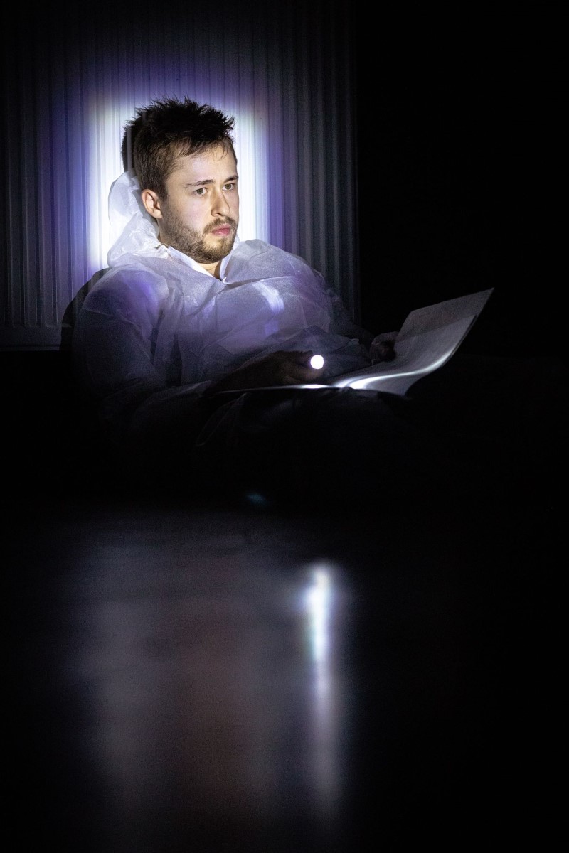 Światło reflektora pada na twarz głównego odtwórcy roli. Siedzi wsparty o ścianę trzymając w dłoni latarkę i tekst.