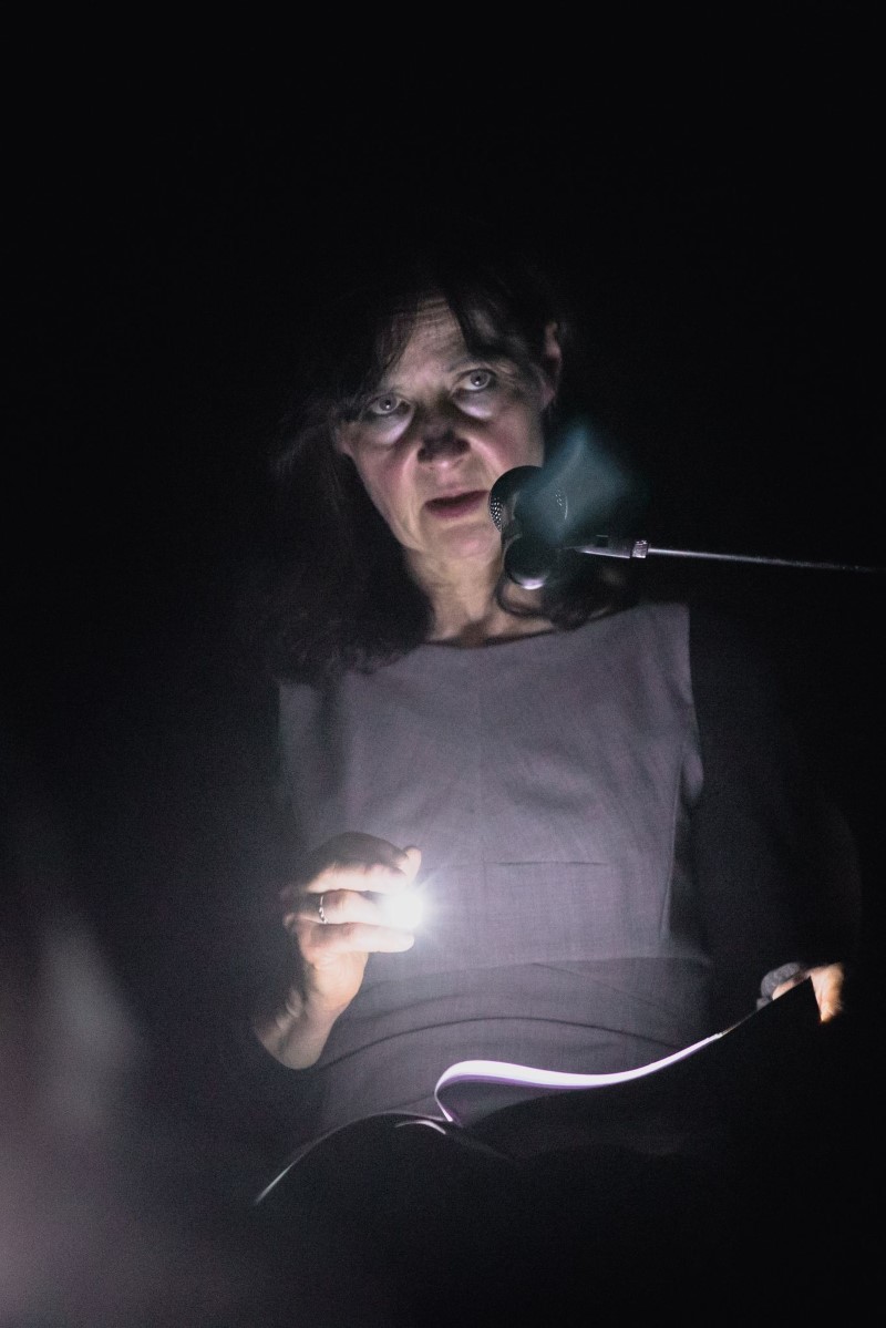 Kobieta w szarej sukience stoi przed mikrofonem, prezentując wyznaczoną partię tekstu. Oświetla go małą latarką.