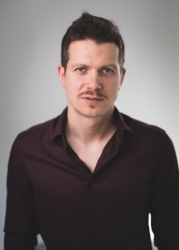 Jakub Mikołajczak, Mężczyzna w granatowej koszulce spogląda w górę; kasztanowe włosy zaczesane na bok.